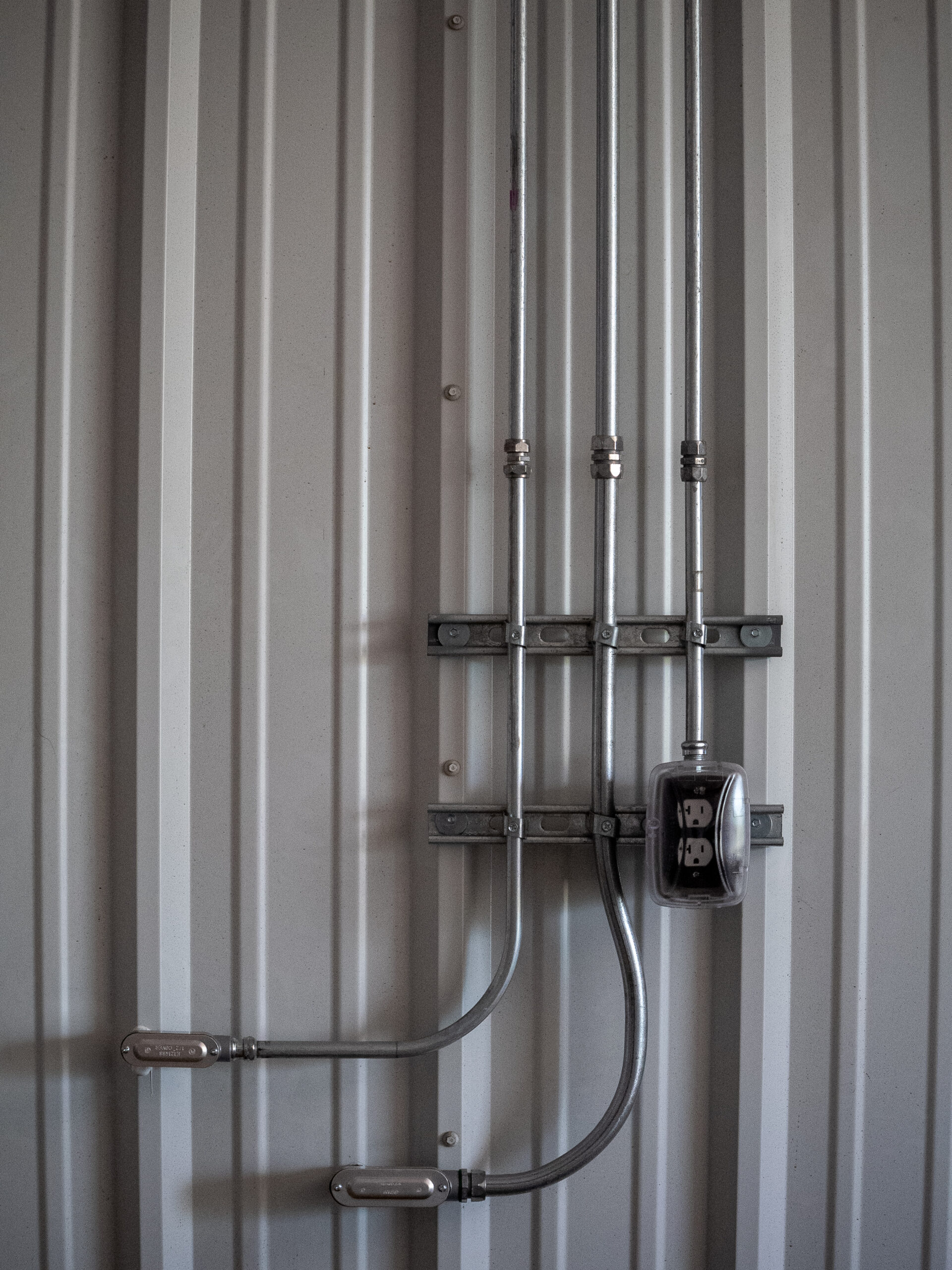 Four metal conduits running vertically along a corrugated metal wall, with a curved conduit at the bottom and an electrical meter mounted on a bracket.